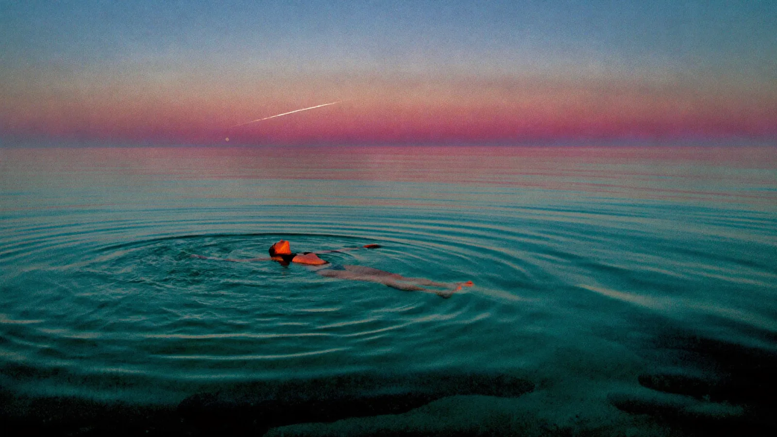 a lone swimmer floating on their back in calm open ocean, in blue hour, a broken moonpath across teal water, a magenta afterglow along the horizon, wide shot, eye level angle, 35mm prime lens, at f/2.8, a single moon source low behind the horizon, no fill, true black in the deep water, palette of teal, magenta, black, still mood, in a style described as "photographic, real film grain, no digital smoothing, the camera at water level"