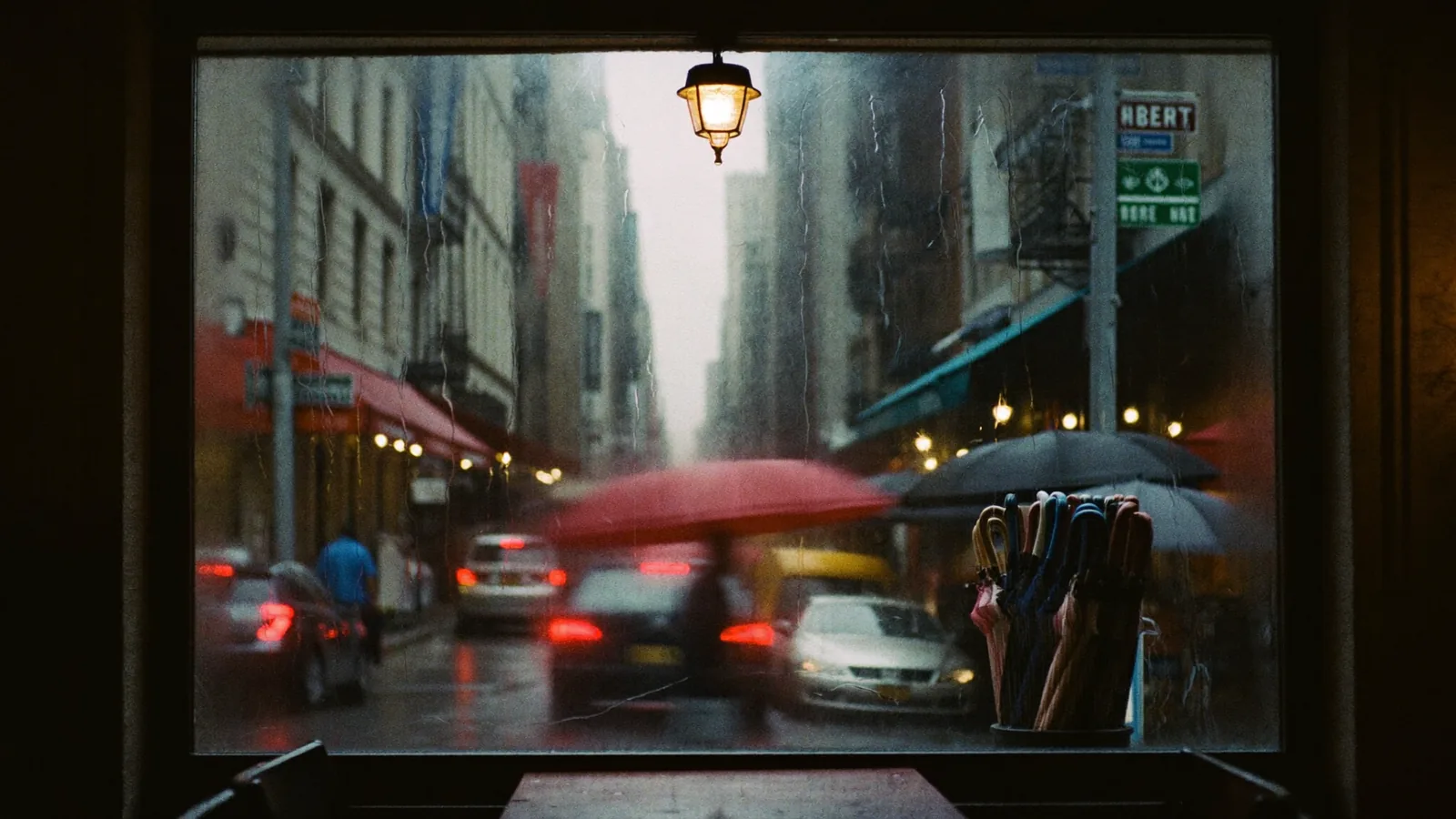a city street seen through a rain-fogged cafe window, one red umbrella crossing, in a street of awnings, taillights and stacked umbrellas compressed into layers, condensation and water tracks on the glass, medium shot, eye level angle, 135mm telephoto lens, at f/2, overcast late afternoon, taillights the only warm source, true black in the wet street, palette of red, slate, amber, hushed mood, in a style described as "photographic, real film grain, no digital smoothing, the glass does the grading"